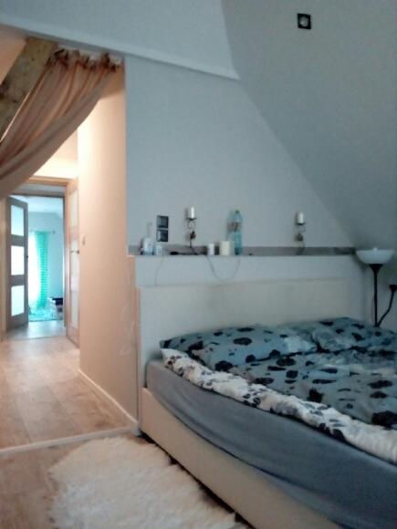 Sypialnia w odcieniach bieli i szarości z łóżkiem z niebieską pościelą w czarne wzory, widok na korytarz z jasną podłogą i otwartymi drzwiami, nad łóżkiem półka z dekoracjami.