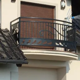 Czarna, metalowa balustrada balkonowa z dekoracyjnymi elementami roślinnymi na tle brązowych rolet okiennych i fragmentu dachu z czarnej dachówki.