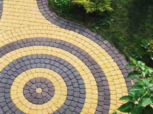 Żółto-szara nawierzchnia ułożona z kostki, tworząca wzór koncentrycznych okręgów, obrzeże z ciemnej kostki, widoczne trawa i krzewy.