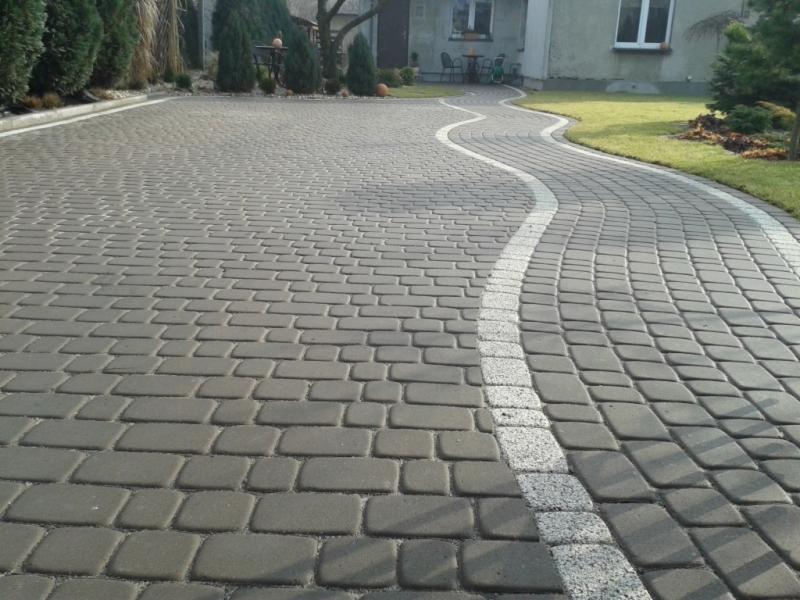 Szara kostka brukowa ułożona w zygzakowaty wzór z białym obramowaniem, prowadząca do domu z ogrodem.