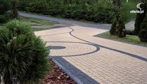 Ułożona kostka brukowa w odcieniach beżu i szarości tworząca wzór na podjeździe, obrzeża z ciemnej kostki, w tle zieleń i drzewa.