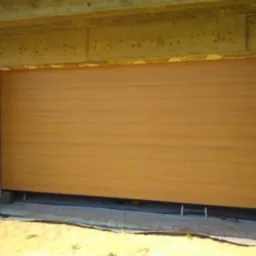 Zamknięta, segmentowa brama garażowa w kolorze jasnego drewna, zamontowana w surowym otworze budynku z cegły.