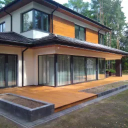 Nowoczesny dom z dużymi, przesuwnymi oknami tarasowymi, drewnianym tarasem i minimalistyczną rabatą przed trawnikiem.