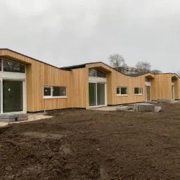 Rząd nowoczesnych domów w trakcie budowy, z drewnianą elewacją i dużymi oknami, widoczne palety z materiałami budowlanymi na nieprzygotowanym terenie.