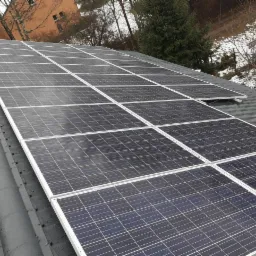 Panel słoneczny na szarym dachu z widocznym fragmentem budynku w tle, pochmurny dzień.