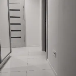 Jasny korytarz z białymi ścianami, białymi płytkami podłogowymi, szarymi drzwiami z czarnymi paskami i lustrzaną szafą wnękową.