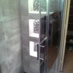 Wykończona kabina prysznicowa z szarymi płytkami i wnękami z mozaiką, widoczny odpływ liniowy i metalowe elementy kabiny.