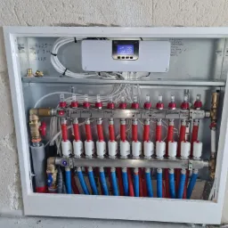 Rozdzielacz ogrzewania podłogowego w metalowej szafce z widocznymi rurami w kolorze czerwonym i niebieskim, z elektronicznym sterownikiem na górze i notatkami odręcznymi na ściankach szafki.