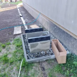 Dwa betonowe elementy z czarnymi pokrywami w betonowej ramie wypełnionej kamieniami, obok ściany budynku z folią kubełkową, na trawniku z niebieskim wężem i kartonowym pudłem.