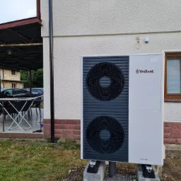 Biała pompa ciepła Vaillant aroTHERM, zamontowana na betonowych bloczkach przed budynkiem z oknami w brązowych ramach, widok z boku.