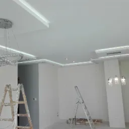 Jasne wnętrze z dekoracyjnym oświetleniem sufitowym, widoczne dwie drabiny malarskie, sufit z zabudową karton-gips i punktowym oświetleniem.