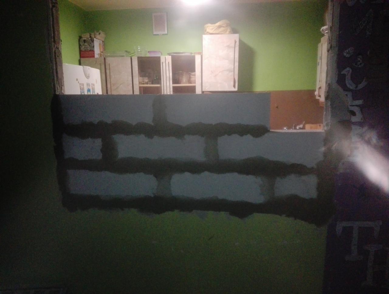 Wznoszenie ścianki działowej z bloczków, widoczne spoiny między bloczkami, zielona ściana w tle i fragmenty wyposażenia kuchni.