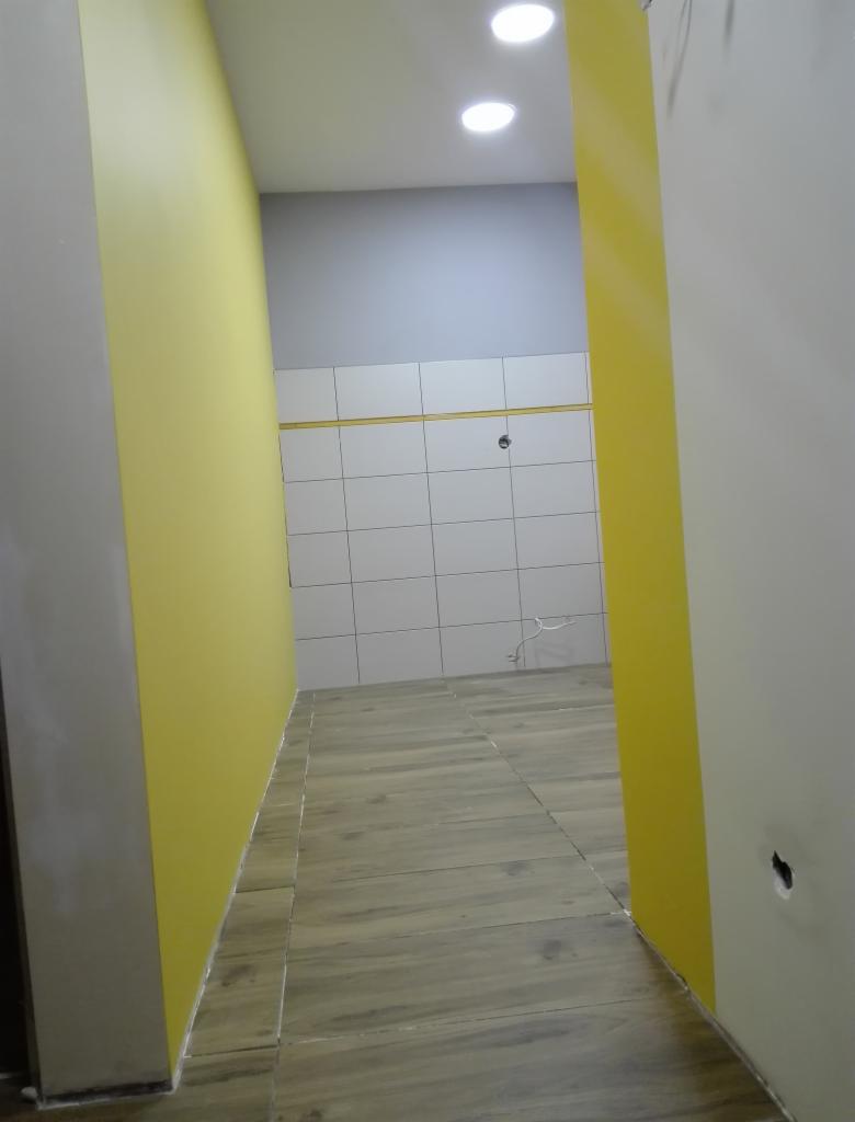 Wnętrze łazienki w trakcie wykańczania: żółta ściana kontrastuje z szarą górną częścią ściany i białymi płytkami, drewnopodobna podłoga, widoczne oświetlenie sufitowe.