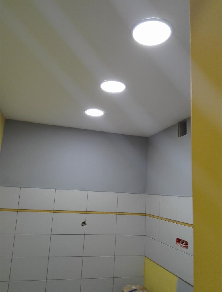 Wykończona łazienka z białymi kwadratowymi płytkami, szarą górną częścią ścian, żółtymi akcentami i trzema okrągłymi lampami sufitowymi