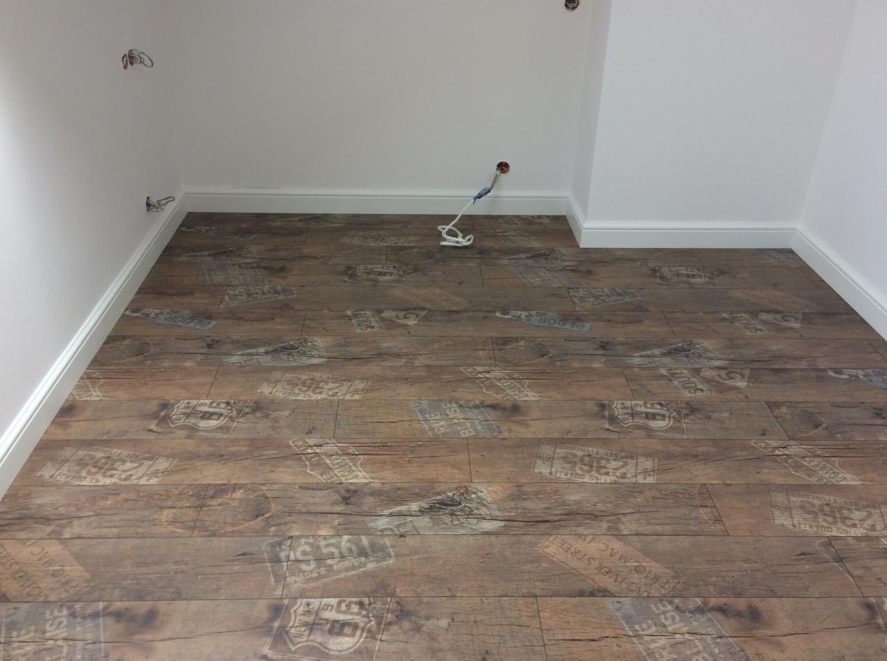 Ukończona podłoga z paneli imitujących stare drewno z motywem tablic rejestracyjnych, widoczne białe listwy przypodłogowe i wystający kabel elektryczny.