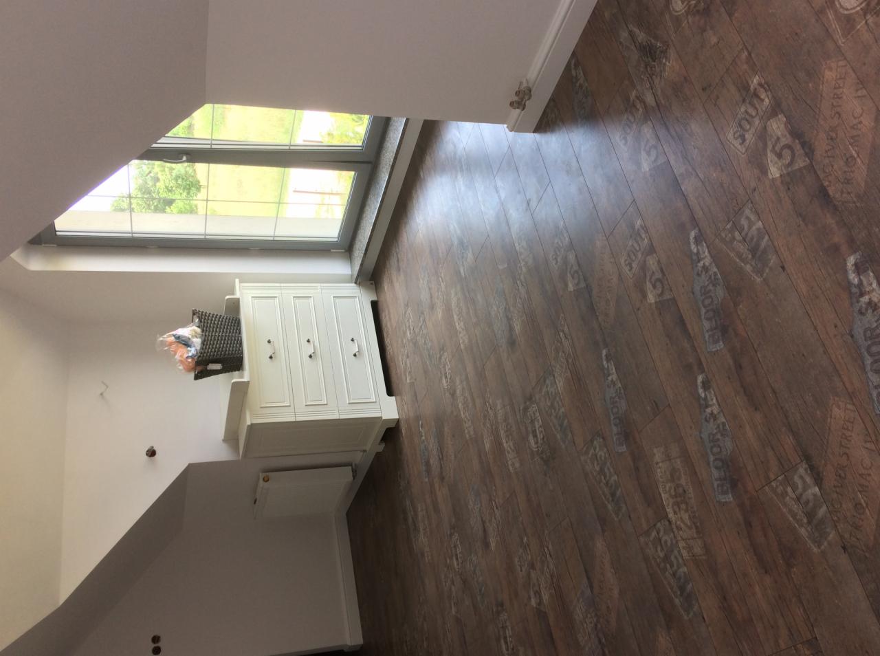 Ukończona podłoga z paneli imitujących drewno z nadrukowanymi napisami, widok na białą komodę pod oknem na poddaszu.