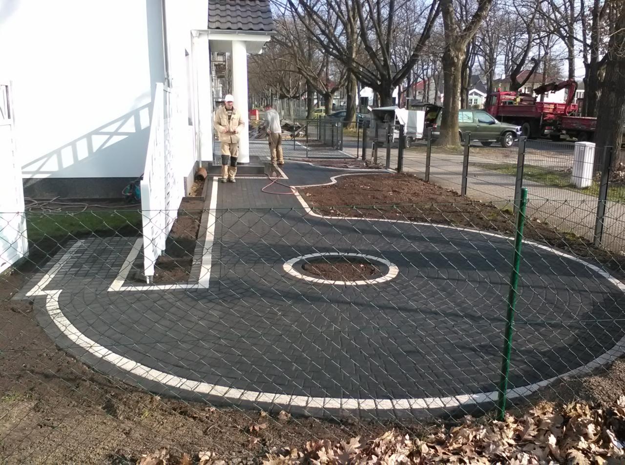 Nowo ułożona, okrągła nawierzchnia z ciemnej kostki z białym obramowaniem, otoczona ziemią przygotowaną pod trawnik, widoczni dwaj pracownicy w oddali i fragment budynku.