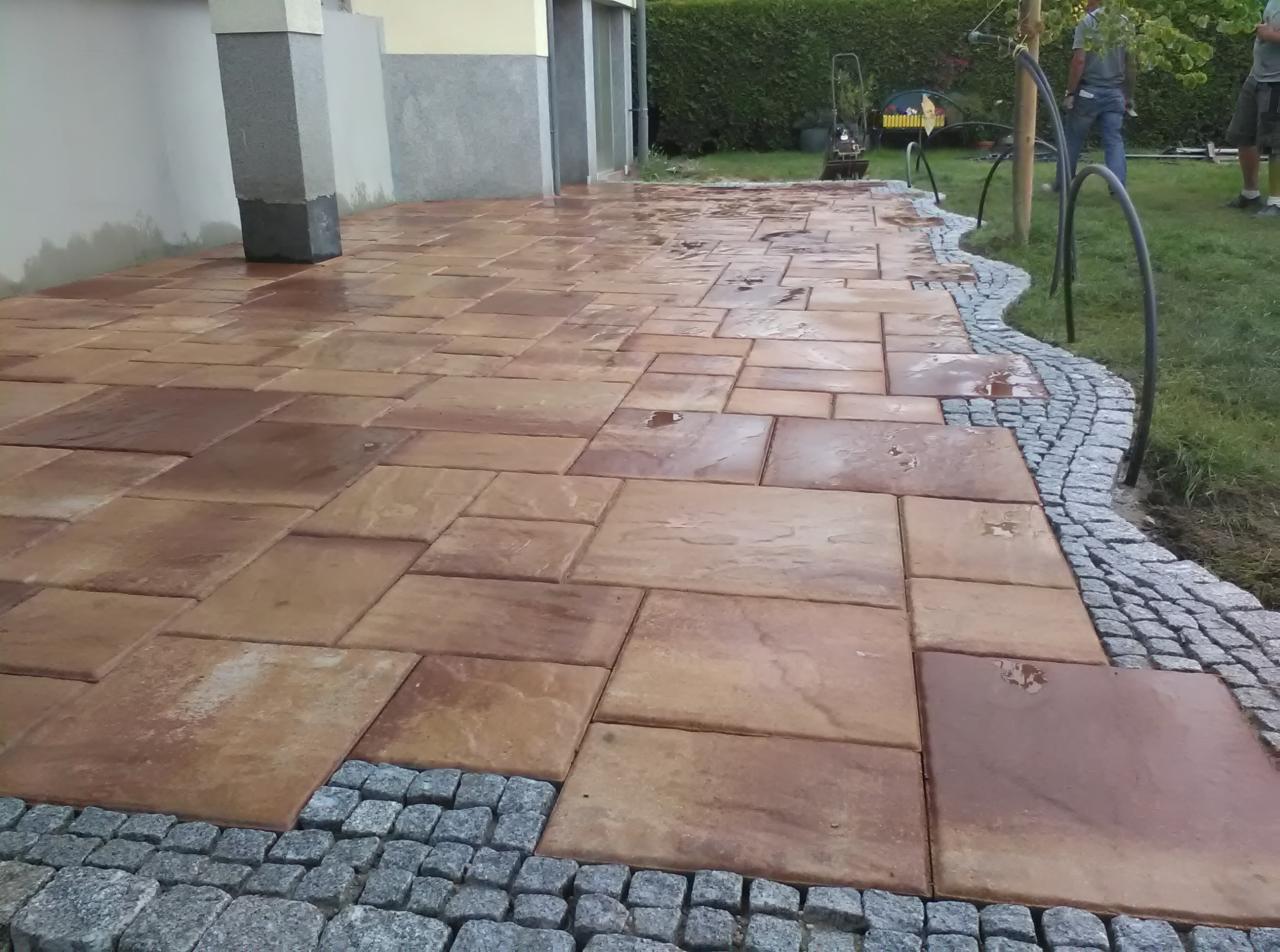 Ułożona nawierzchnia z dużych, prostokątnych płyt w odcieniach brązu, obramowana szarą kostką granitową, widok na budynek w tle.