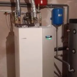 Nowoczesna, biało-szara pompa ciepła z podłączonymi rurami, zbiornikami w kolorze czerwonym i niebieskim, na tle surowej ściany w trakcie remontu.