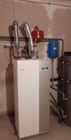 Nowoczesna, biało-szara pompa ciepła z podłączonymi rurami, zbiornikami w kolorze czerwonym i niebieskim, na tle surowej ściany w trakcie remontu.