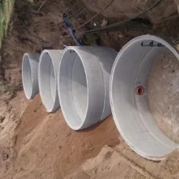 Betonowe kręgi studni ułożone w wykopie, widoczny fragment koparki i krajobraz wiejski w tle.