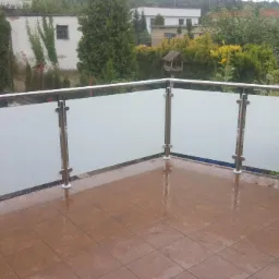 Balkon z nowoczesną, matową, szklaną osłoną z nierdzewnymi słupkami, widok na zieleń ogrodu i budynki w tle, mokra, brązowa podłoga z płytek.