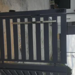 Nowoczesna, metalowa brama posesyjna z poziomymi szczeblami, widoczny zamek elektroniczny i uchwyt ze stali nierdzewnej, na tle domu jednorodzinnego i pochmurnego nieba.