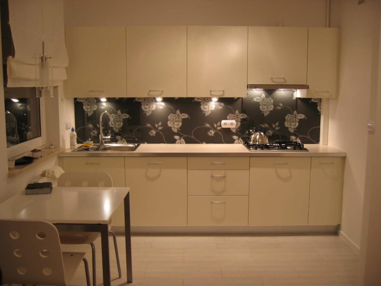 Jasna kuchnia z kremowymi szafkami, czarnym panelem ściennym w białe kwiaty, oświetleniem podszafkowym i stołem z krzesłami.