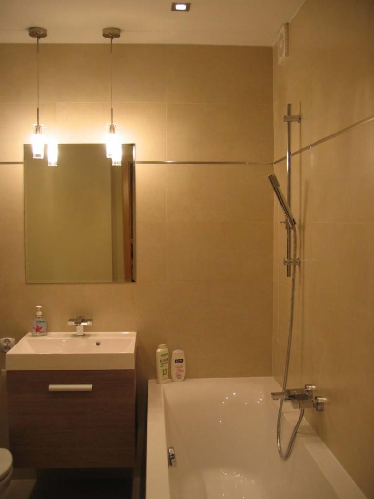 Jasna łazienka z beżowymi płytkami, umywalką z szafką w kolorze drewna, wanną i minimalistycznym oświetleniem punktowym nad lustrem.