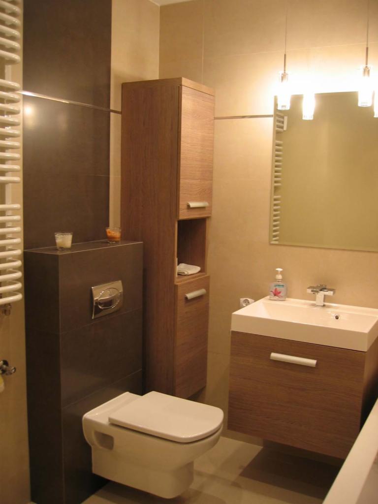 Nowoczesna łazienka z wiszącą toaletą w zabudowie z ciemnych płytek, drewnianą szafką łazienkową z umywalką nablatową oraz wysoką szafką w kolorze drewna, oświetlenie punktowe nad lustrem.