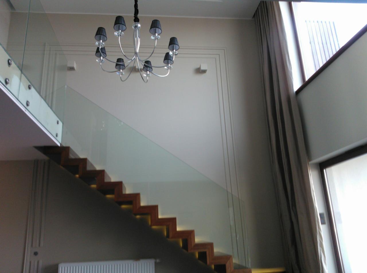 Schody drewniane z podświetleniem LED, szklana balustrada, nowoczesny żyrandol z czarnymi abażurami, wysokie okno z zasłonami w jasnym wnętrzu.