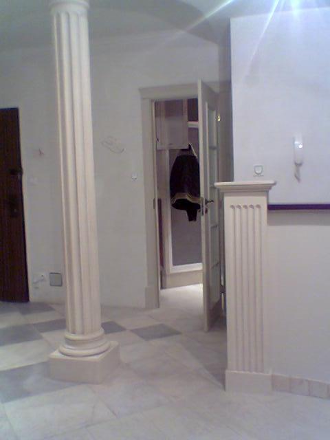 Wnętrze z marmurową podłogą w szachownicę, białą kolumną z żłobieniami i otwartymi drzwiami z widocznym wieszakiem na ubrania.