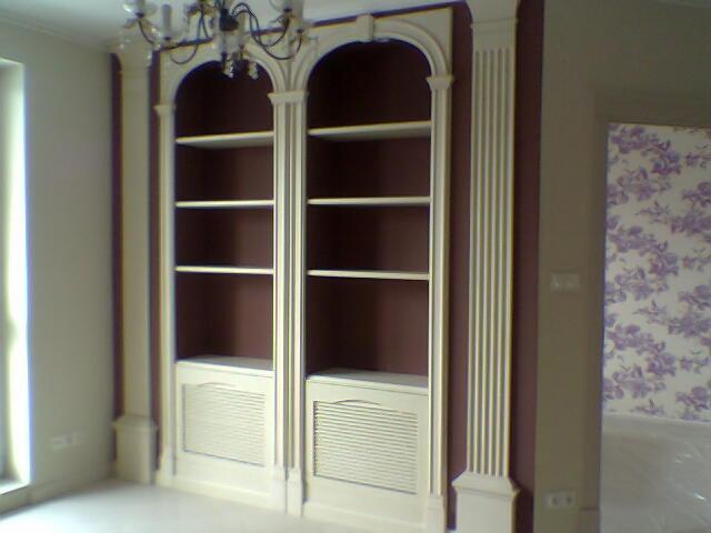 Dwa białe, wbudowane regały z półkami i dekoracyjnymi kolumnami, wnętrze pomalowane na burgundowo, w tle pokój z fioletową tapetą w kwiaty, widoczny żyrandol.