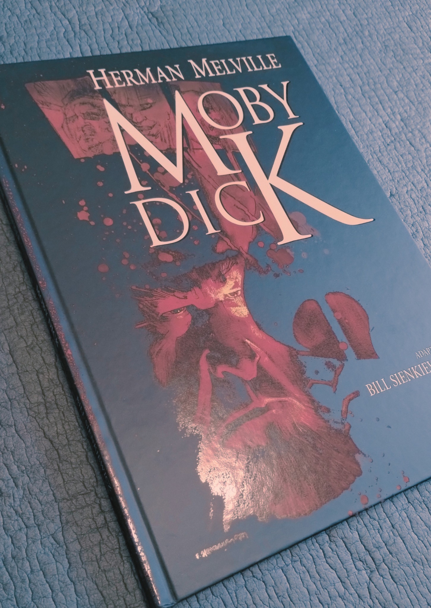 Książka 'Moby Dick' Hermana Melville'a z okładką autorstwa Billa Sienkiewicza, leżąca na teksturowanym, niebieskim tle.
