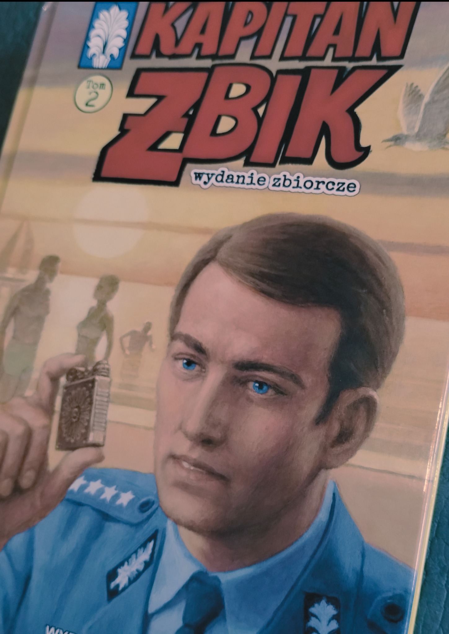 Okładka książki 'Kapitan Żbik, wydanie zbiorcze, Tom 2' z ilustracją portretową kapitana w mundurze trzymającego zapalniczkę na tle plaży z ludźmi.