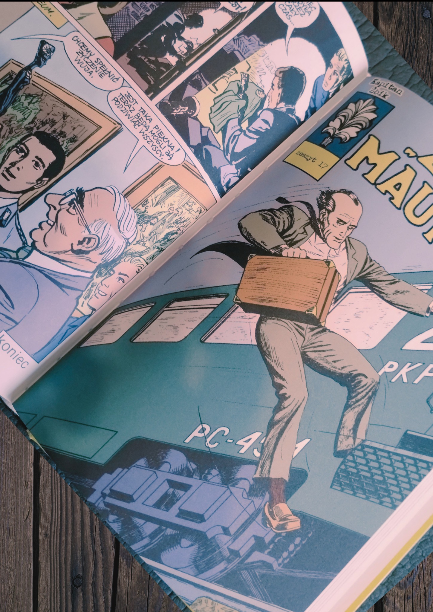 Otwarty komiks na drewnianym stole, prezentujący ilustracje w stylu retro z dialogami w dymkach i sceną z mężczyzną z walizką wychodzącym z pociągu.