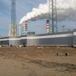 Nowoczesny budynek przemysłowy obłożony płytami warstwowymi w kolorze szarym i białym, kominy elektrowni w tle, widoczne słupy wysokiego napięcia.