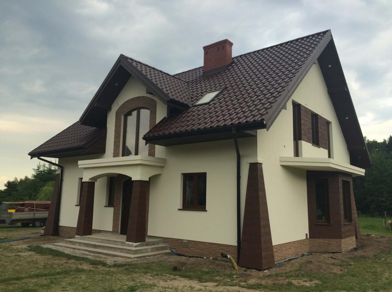 Dwukondygnacyjny dom z beżową elewacją, brązową dachówką i elementami dekoracyjnymi imitującymi kamień, widoczny w pochmurny dzień.