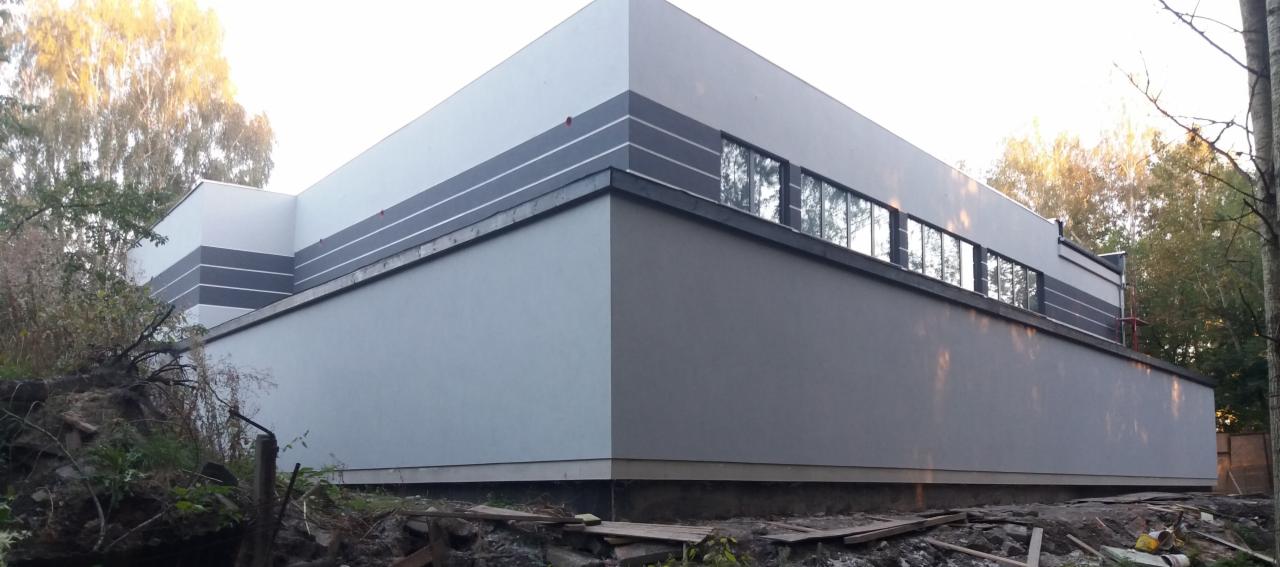 Elewacja budynku po remoncie, z szarym tynkiem i białymi pasami na górze, widoczne okna z refleksami drzew.