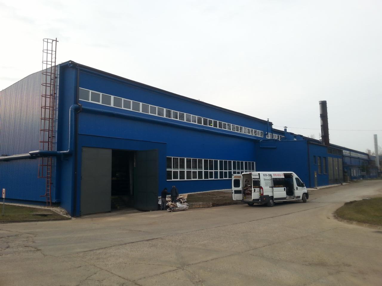 Niebieski budynek przemysłowy z blachy trapezowej, widoczna drabina przeciwpożarowa, otwarte wrota i zaparkowany bus z reklamą firmy elewacyjnej.