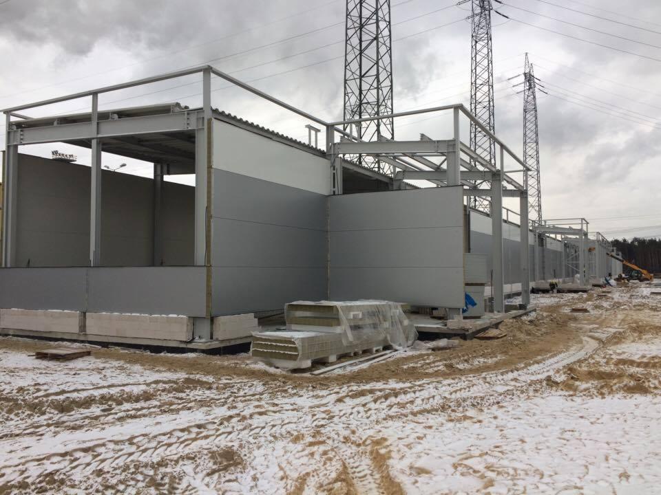 Budowa hali z płyt warstwowych w zimowej scenerii, widoczne elementy konstrukcyjne i przygotowane płyty, w tle słupy wysokiego napięcia.