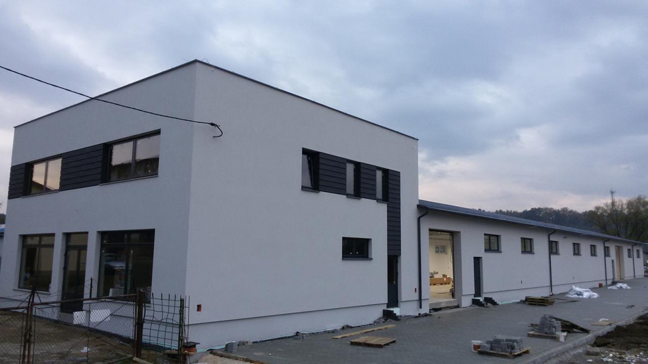 Nowoczesny budynek biurowy z jasną elewacją, ciemnymi akcentami wokół okien i widocznymi materiałami budowlanymi na placu przed budynkiem.