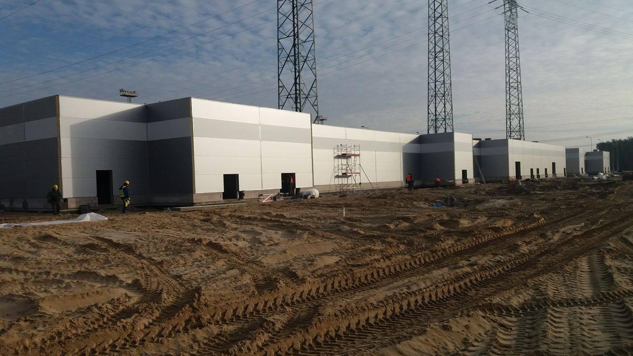 Montaż hali z płyt warstwowych: widok na budynek w trakcie budowy, z widocznymi pracownikami w żółtych kaskach i rusztowaniem, oraz z ziemią z wyraźnymi śladami opon.