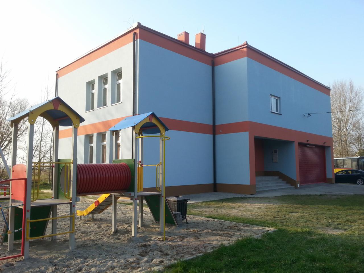 Budynek ocieplony z placem zabaw na pierwszym planie; pastelowe kolory elewacji z pomarańczowymi akcentami, widoczne okna i brama garażowa.