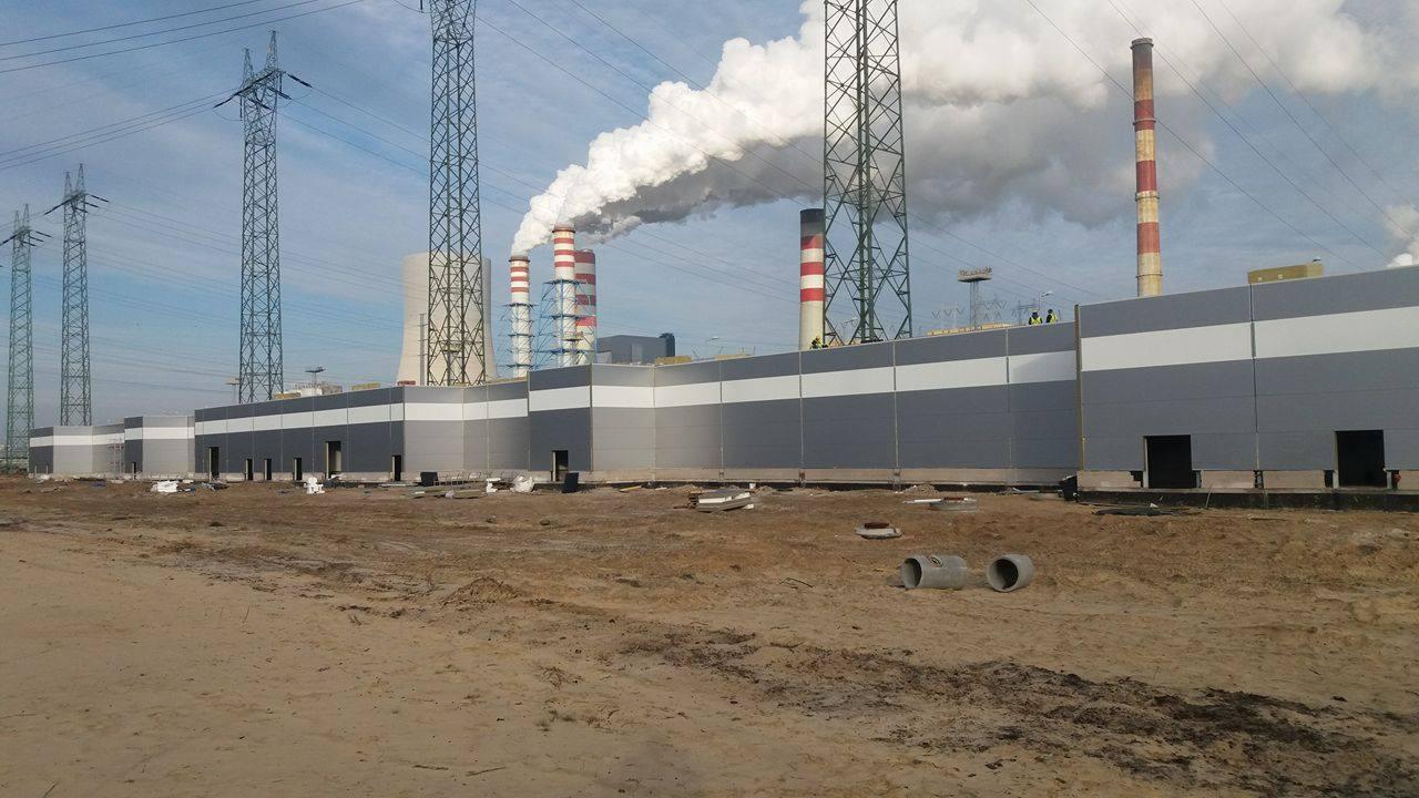 Nowoczesny budynek przemysłowy obłożony płytami warstwowymi w kolorze szarym i białym, kominy elektrowni w tle, widoczne słupy wysokiego napięcia.