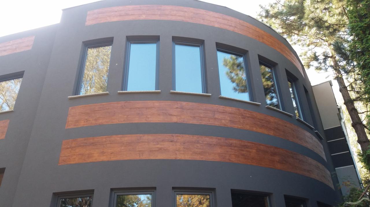 Nowoczesna elewacja budynku z szarym tynkiem i dekoracyjnymi pasami imitującymi drewno, widoczne okna z refleksami drzew.