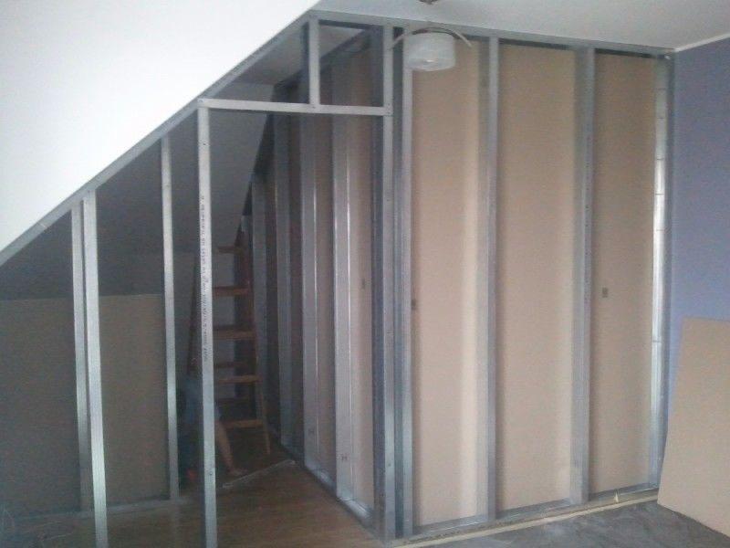 Konstrukcja ścian działowych z profili stalowych i płyt gipsowo-kartonowych w pomieszczeniu ze skosem, widoczna drewniana drabina w tle.