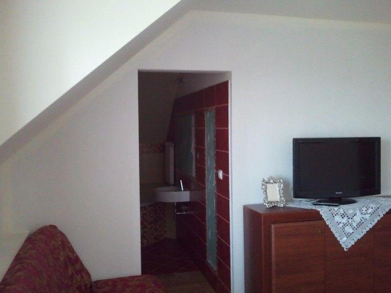 Pokój na poddaszu z widokiem na łazienkę z czerwonymi płytkami i umywalką, telewizor na komodzie z białą serwetką