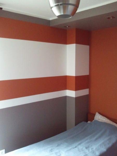 Pokój z łóżkiem, ściany pomalowane w poziome pasy: pomarańczowy, biały i szary, lampa sufitowa w stylu industrialnym.