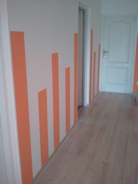 Korytarz z pomalowanymi ścianami w pionowe, nieregularne pasy w odcieniu pomarańczowym, parkiet na podłodze, białe drzwi na końcu korytarza.